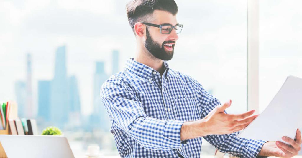 Ein Mann mit braunen, kurzen Haaren, einem Bart und einer Brille hält ein Dokument in der Hand, lächelt und freut sich über Leichte Sprache