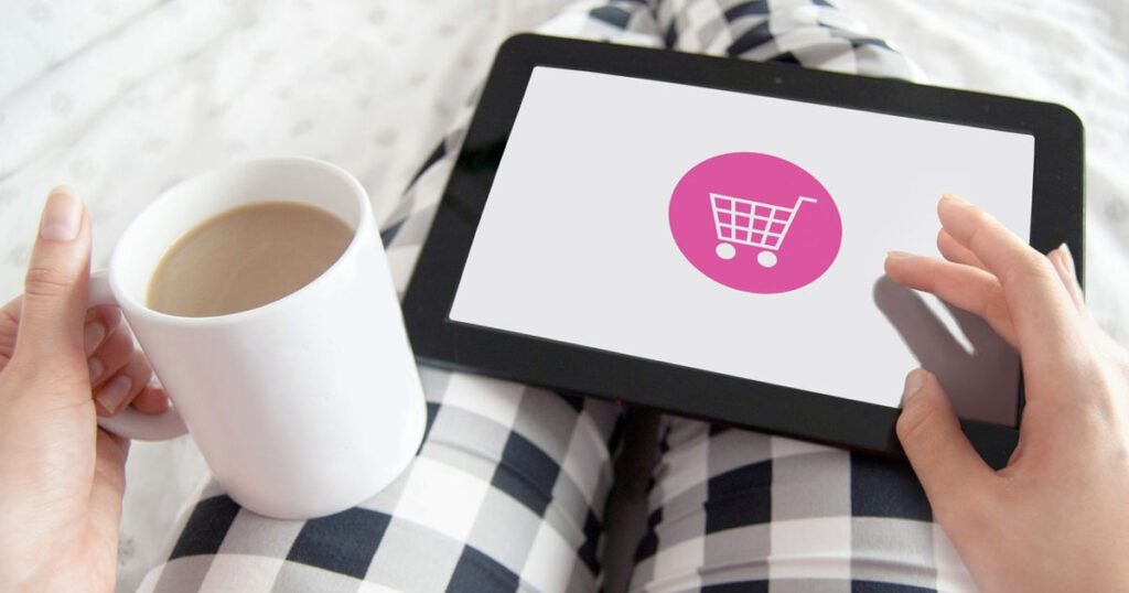 Eine Frau sitzt auf dem Sofa und kauft mit einem Tablet Online ein. In der linken Hand hält Sie eine Tasse Kaffee. Für Sie ist der Onlineshop barrierefrei!