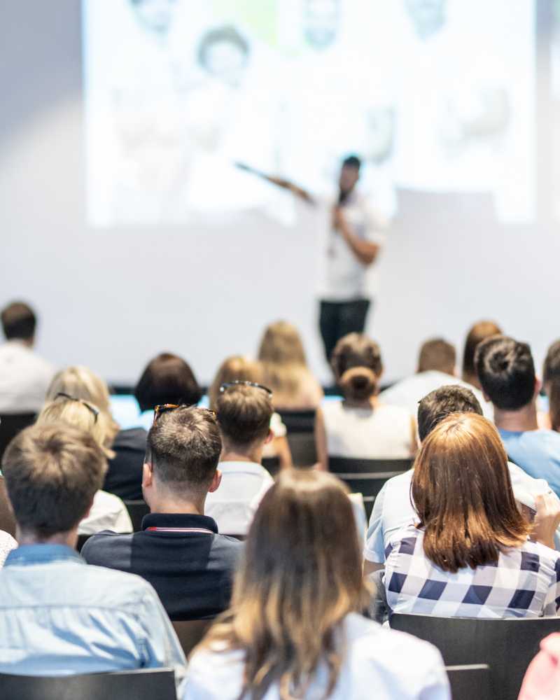 Im Hintergrund steht eine Person mit Mikrofon auf der Bühne. Die Person ist unscharf dargestellt. Der Fokus ist auf die Hinterköpfe des Publikums gerichtet.