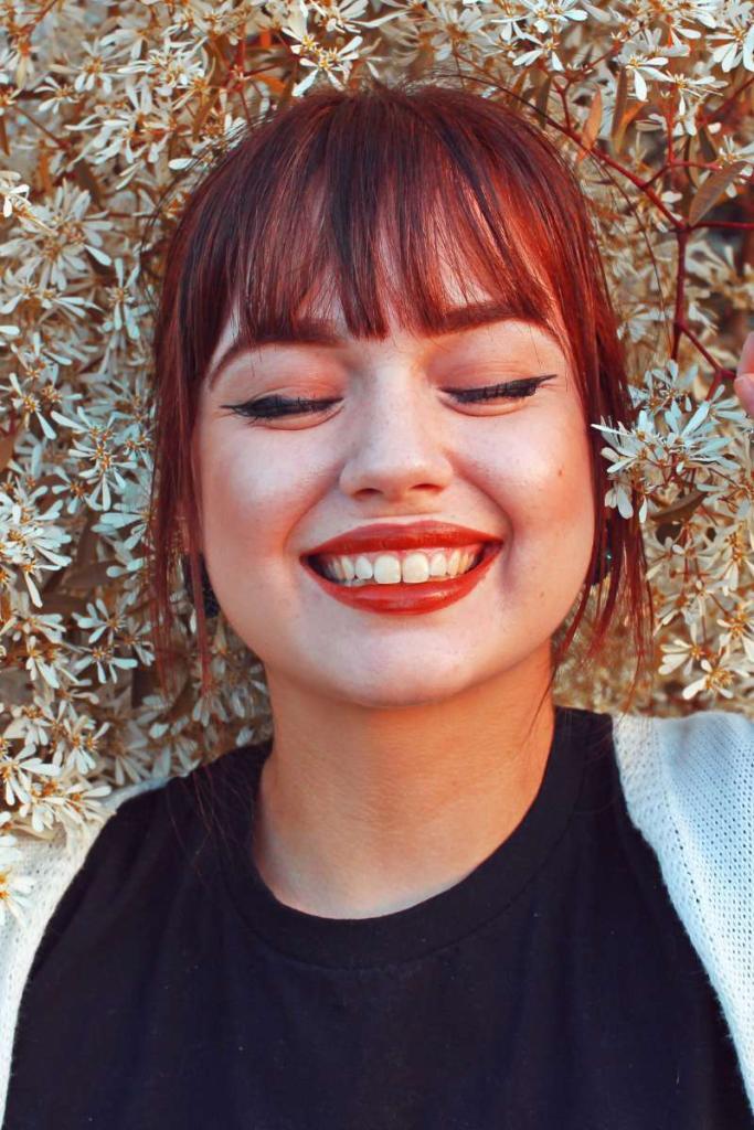Eine Frau mit roten Haaren und einem roten Lippenstift hat geschlossene Augen und lächelt mit zufriedenem Blick.