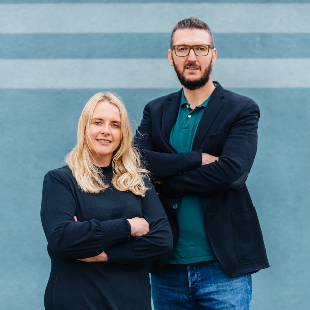 Eine Frau und ein Mann stehen nebeneinander vor einer blauen Wand mit zwei horizontalen Streifen. Die Frau trägt ein schwarzes Kleid und hat blonde, schulterlange Haare. Der Mann trägt eine Brille, ein grünes Polohemd, ein dunkles Sakko und eine Jeans. Beide schauen in die Kamera und lächeln leicht.