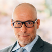 Ein Mann mit kurzen Haaren, einem dunklen Anzug und einer hellen Krawatte mit hellem Hemd