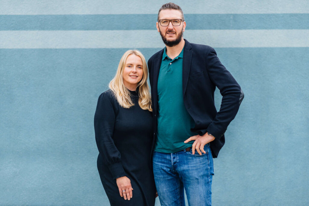 Eine Frau und ein Mann stehen nebeneinander vor einer blauen Wand mit zwei horizontalen Streifen. Die Frau trägt ein schwarzes Kleid und hat blonde, schulterlange Haare. Der Mann trägt eine Brille, ein grünes Polohemd, ein dunkles Sakko und eine Jeans. Beide schauen in die Kamera und lächeln leicht.