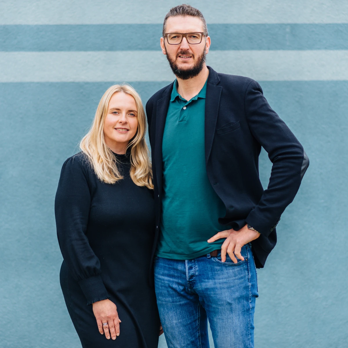 Eine Frau und ein Mann stehen nebeneinander vor einer blauen Wand mit zwei horizontalen Streifen. Die Frau trägt ein schwarzes Kleid und hat blonde, schulterlange Haare. Der Mann trägt eine Brille, ein grünes Polohemd, ein dunkles Sakko und eine Jeans. Beide schauen in die Kamera und lächeln leicht.